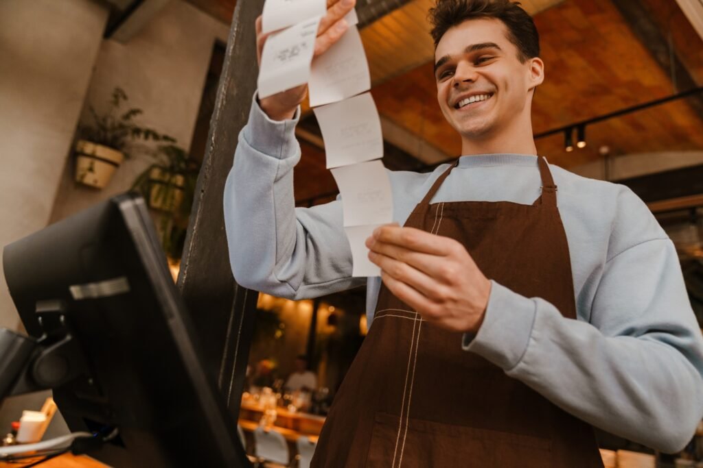 Young waiter man standing in front of computer and holding receipt
